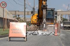 #ParaQuemNaoPodeVer A foto exibe uma obra na rua. Na imagem, há uma placa de aviso com o texto "ATENÇÃO OBRAS À FRENTE". Ao fundo, é possível ver uma escavadeira amarela e uma cerca de proteção laranja. No canto superior direito, há uma placa de trânsito indicando o limite de velocidade de 30 km/h. A cena parece estar em uma área urbana com calçadas e postes de eletricidade.