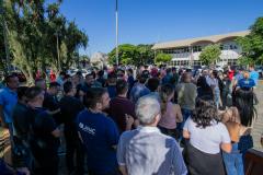 #ParaQuemNaoPodeVer A foto mostra um grupo grande de pessoas reunidas ao ar livre, em um local que parece ser uma área urbana com árvores e um prédio ao fundo. As pessoas estão em fila ou aglomeradas, e o céu está claro e azul.