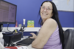 #praquemnaopodever Uma mulher de cabelos negros e lisos, regata lilas e óculos sorri para a camera. Ela está sentada em frente ao computador, mas precisou virar de lado para posar pra foto. O ambiente é de escritório.