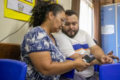 #praquemnaopodever uma mulher negra de blusa branca com plantas azuis mexe no celular ao lado de um rapaz de camisa branca com listras azul e laranja. Os dois estão sentados dentro de uma sala de aula e olham para o aparelho, enquanto a senhora de meia idade e óculos digita no celular.