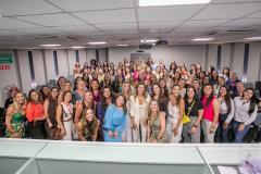 #ParaQuemNaoPodeVer Imagem mostra um grande grupo de pessoas, principalmente mulheres, posando juntas em um ambiente interno, possivelmente uma sala de conferências ou auditório. As pessoas estão sorrindo e parecem estar participando de um evento ou reunião. Algumas estão vestindo roupas coloridas e outras em tons mais neutros. 