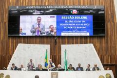 #praquemnaopodever 10 pessoas estão sentadas em um púlpito alto e com fachada branca. Em pé está um homem de terno cinza e gravata rosa, segurando um microfone na mão e discursando enquanto gesticula com a mão direita. Seu perfil aparece no telão ao fundo, ao lado de um texto com os dizeres "sessão solene em homenagem aos 90 anos do Portos Paraná"