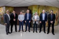 #ParaQuemNaoPodeVer Imagem exibe uma imagem de seis homens em pé, lado a lado, em um ambiente interno. Eles estão vestidos formalmente, com ternos e camisas sociais. Ao fundo, há uma parede com uma pintura colorida. 