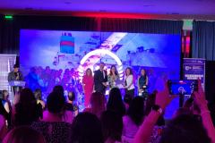 #ParaQuemNaoPodeVer A imagem mostra um evento. Há várias pessoas no palco, algumas segurando microfones, e outras na plateia, algumas tirando fotos com celulares. O ambiente está iluminado com luzes roxas e azuis. No fundo, há um painel com logotipos e textos que não são legíveis nesta imagem.