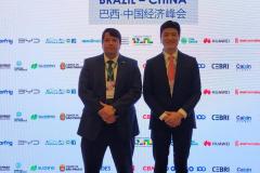 #ParaQuemNaoPodeVer A imagem mostra dois homens de terno em pé, lado a lado, em um evento. O fundo tem um banner com logotipos de várias empresas e as palavras "SUMMIT Valor Econômico BRAZIL-CHINA". Há também caracteres chineses no banner. Os homens estão de frente para a câmera, um deles segurando um microfone.