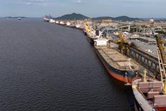 #ParaQuemNaoPodeVer A imagem exibe um porto com um grande navio atracado. O navio está carregado com contêineres e há guindastes ao redor. A água do mar é visível à esquerda, e ao fundo, há uma cidade com colinas.