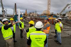 #praquemnaopodever homens e mulheres de colete verde limão estão em uma obra olhando para um rapaz de capacete branco que aponta ao longe. O céu está cinza, o chão de barro está molhado. Ao fundo, máquinas estão suspensas por causa da chuva que dá sinais de que irá voltar