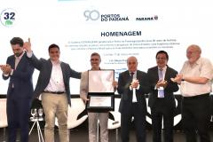 #praquemnaopodever homem te terno cinza e óculos de grau segura uma caixa aberta com uma placa dentro.  Ele e outros cinco homens brancos de roupa social estão em cima de um palco, alguns sorriem, outroas fazem joinha. Ao fundo, aparece escrito "Homenagem 90 anos" no telão