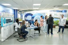 #ParaQuemNaoPodeVer Um grupo de pessoas está em um ambiente interno que parece ser uma feira ou evento de negócios. Há uma mesa com duas mulheres sentadas, uma delas usando jaleco branco e a outra com cabelo comprido e loiro. Elas estão interagindo com outras pessoas em pé. No fundo, há um banner com o texto "Portos do Paraná" e imagens de paisagens. Algumas pessoas estão vestindo jalecos brancos, sugerindo que podem ser profissionais de saúde ou de outra área técnica. O ambiente é bem iluminado, com teto d