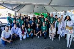 #ParaQuemNaoPodever Um grupo grande de pessoas está posando para uma foto em um ambiente externo. A maioria delas está usando camisetas verdes com a inscrição "ABRIL VERDE 2025". Algumas pessoas estão agachadas na frente, enquanto outras estão em pé atrás. Há uma tenda ao lado direito da imagem e algumas bexigas verdes. No canto inferior esquerdo, há uma mesa branca com alguns objetos em cima, incluindo um livro amarelo e um panfleto azul.