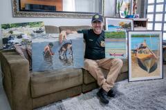 #ParaQuemNaoPodeVer Homem sentado em um sofá segurando duas pinturas. Ele está usando uma camiseta preta e um boné azul com um emblema. As pinturas mostram cenas de praia: uma com duas pessoas e um cachorro na água, e outra com um barco na areia. Ao lado dele, há mais pinturas e uma moldura decorativa na parede. O ambiente parece ser uma sala de estar, com um sofá bege e uma mesa com objetos ao fundo.