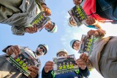 #ParaQuemNaoPodeVer Imagem exibe várias pessoas em um círculo, olhando para a câmera de baixo para cima. Elas estão segurando panfletos ou revistas que têm a imagem de trabalhadores de construção civil na capa. O texto na capa diz: "INFLUÊNCIA POSITIVA: ESSA É A POSTURA DA PREVENÇÃO". As pessoas na foto estão usando capacetes de segurança e óculos de proteção, sugerindo que são trabalhadores de uma obra ou indústria.