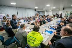 #praquemnaopodever setenta pessoas estão sentadas olhando para um senhor em pé, de camisa azul escura e calça bege. Ele segura um microfone na mão e com olhar focado por trás dos óculos, observa o público que o acompanha. A sala está com as cortinas brancas fechadas e ao fundo a imprensa grava o encontro.