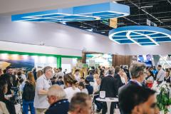 #ParaQuemNaoPodeVer Foto exibe um evento ou feira com muitas pessoas. Há uma estrutura grande e moderna no teto, com formas geométricas azuis. As pessoas estão em pé e algumas estão sentadas em mesas, parecendo interagir ou observar algo. O ambiente está movimentado e há uma variedade de pessoas, algumas com mochilas. 