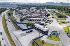 #ParaQuemNaoPodeVer Imagem aérea de um grande pátio industrial ou logístico. No centro da imagem há um prédio principal de formato retangular, com várias vagas de estacionamento ao redor. À direita do prédio, há uma grande quantidade de caminhões ou carretas estacionados em fileiras organizadas. O local é cercado por áreas verdes e ruas asfaltadas, com alguns veículos circulando. Ao fundo, é possível ver uma cidade e montanhas.