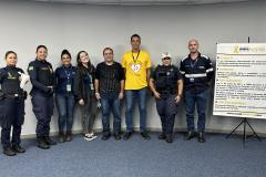 #ParaQuemNaoPodeVer Foto de um grupo de oito pessoas, sendo quatro delas usando uniformes de agentes de trânsito ou policiais, e as outras quatro usando roupas casuais. Eles estão em pé, lado a lado, em frente a uma parede clara e um tapete azul. À frente do grupo, há um banner com informações sobre o movimento Maio Amarelo, que é uma campanha de conscientização para a segurança no trânsito. O banner tem detalhes em amarelo e branco, com o símbolo do laço amarelo. Todos estão sorrindo e olhando para a câmer