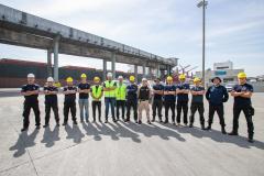 #ParaQuemNaoPodeVer Foto de um grupo de pessoas em um porto. Elas estão alinhadas, usando capacetes de segurança amarelos e brancos, e algumas vestem coletes refletivos. Ao fundo, há uma estrutura de concreto que de carregamento de granéis.