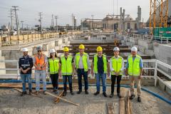 #ParaQuemNaoPodeVer Grupo de oito pessoas em um canteiro de obras. Todos estão usando equipamentos de segurança, como capacetes e coletes refletivos. A maioria veste coletes amarelos, enquanto uma pessoa usa um colete laranja. O cenário ao fundo mostra estruturas de concreto e uma grua amarela, indicando que estão em uma área de construção. O céu está nublado e há postes de eletricidade ao fundo.