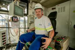 #ParaQuemNaoPodeVer Foto de um homem sentado em uma cabine de operação. Ele está usando capacete branco com o logotipo da empresa "ICTSI" e uniforme azul com faixas refletivas amarelas. O homem está sentado em uma cadeira diante de um painel de controle com vários botões coloridos e alavancas. À frente dele, há uma tela de computador com gráficos e informações técnicas. A cabine tem janelas grandes, permitindo ver estruturas metálicas e o que parece ser um terminal portuário ao fundo. O ambiente sugere que 