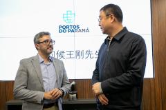 #praquemnaopodever na foto, dois homens conversam em frente a uma tela branca com logo escrita "Portos do Paraná" e dizeres em mandarim embaixo. A esquerda, o homem branco de terno cinza, camisa azul e óculos segura as mãos enquanto conversa com um homem chinês mais alto, de óculos, camisa e calça pretas.
