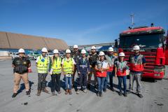 #ParaQuemNaoPodeVer Imagem mostra grupo de pessoas posando em frente a um caminhão vermelho. Todos estão usando capacetes de segurança e coletes refletivos, indicando que podem estar em um ambiente de trabalho ou construção. Ao fundo, há um grande galpão e o céu está azul.