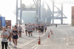 #praquemnaopodever foto mostra multidao de atletas atravessando o cais de um porto correndo. Ao fundo, a neblina encobre metade de um navio. Os guindastes gigantes azuis estão ao lado da embarcação, mas sem se moverem.