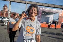 #praquemnaopodever na foto, ao centro, idosa com camisa cinza e letras em laranja escrito 90 Portos do Paraná, ergue com um sorriso no rosto a medalha recebida após concluir a Corrida do Porto.