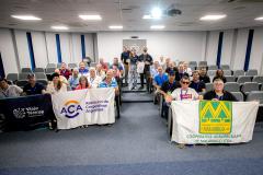 #ParaQuemNaoPodeVer Foto de um grupo de pessoas sentadas em cadeiras de auditório, posando para a câmera. Elas estão sorrindo e segurando três faixas diferentes. A faixa do topo, à esquerda, tem o texto “Viajes Teóricos” e “Colectivo”, a faixa do meio tem o logotipo da ACA e diz “Asociación de Cooperativas Argentinas”, e a faixa da frente, em destaque, tem um grande “M” amarelo e verde e o texto “Cooperativa Agropecuária de Malabrigo Ltda.”. O ambiente parece ser uma sala de reuniões ou auditório, com cadei