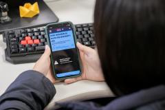 #ParaQuemNaoPodeVer Imagem mostra uma foto de uma pessoa segurando um celular. O celular está exibindo uma página da internet com o título “COMO FUNCIONAM OS GOLPES POR WHATSAPP E SMS”. Abaixo do título, há um texto menor, mas não está totalmente legível na imagem. O celular está em cima de um teclado de computador com algumas teclas vermelhas e outras pretas. No canto superior direito da mesa, há um objeto amarelo que parece ser uma dobradura de papel (origami). A pessoa está usando uma blus