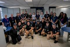 #ParaQuemNaoPodeVer Foto de grupo em ambiente de escritório. Aproximadamente vinte pessoas estão reunidas, a maioria usando camisetas pretas com logotipos e calças bege, sugerindo que fazem parte de uma equipe ou evento. Algumas pessoas estão de crachá e há diversidade de gênero e idade. Todos estão sorrindo e posando para a foto, alguns agachados na frente e outros em pé atrás. Ao fundo, há divisórias de escritório, mesas e cadeiras.