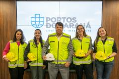#ParaQuemNaoPodeVer Foto de cinco pessoas em pé, todas usando coletes de segurança amarelo e segurando capacetes (a maioria amarelos, um branco). Elas estão sorrindo e posando para a foto em frente a um painel com o logotipo azul e as palavras “PORTOS DO PARANÁ LOGÍSTICA INTELIGENTE”. O grupo é composto por quatro mulheres e um homem, todos com aparência profissional.
