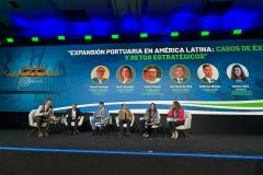 #ParaQuemNaoPodeVer Palco de conferência com seis pessoas sentadas em cadeiras, participando de um painel de discussão. Ao fundo, há um grande painel azul com a imagem de um contêiner portuário e o título do evento em espanhol: "Expansión portuaria en América Latina: casos de éxito y retos estratégicos". Abaixo do título, aparecem fotos e nomes dos participantes do painel, com seus respectivos cargos e empresas. O ambiente é formal, com todos vestidos de maneira profissional. O palco está bem iluminado e há