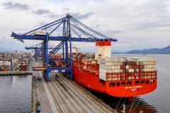 #ParaQuemNaoPodeVer Imagem de um navio cargueiro vermelho atracado em um porto. O navio está carregado com vários contêineres empilhados e há guindastes azuis grandes posicionados para carregar ou descarregar os contêineres. À esquerda, é possível ver parte do cais com caminhões e veículos pequenos. O mar está calmo e o céu está parcialmente nublado.