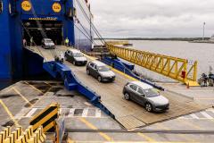 #ParaQuemNaoPodeVer A imagem mostra uma rampa amarela de acesso à balsa, que está ancorada em um porto. Há cinco carros visíveis, alinhados e entrando na balsa, que é azul. No fundo, vê-se o rio e parte da margem oposta. Sinais circulares amarelos na entrada da balsa indicam altura máxima de 7,5 metros e largura máxima de 5,2 metros. O ambiente parece ser de um terminal portuário.