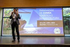 #ParaQuemNaoPodeVer Foto de um homem em pé, falando ao microfone em um palco. Ele está usando calça preta, sapato preto, camisa clara e blazer escuro. Atrás dele há um telão grande com a seguinte mensagem:  "PARANAENSE DE COMEX LOGÍSTICA E INFRAESTRUTURA 24 e 25 JUN 2025 ISUPAR, PARANAGUÁ-PR"  No canto inferior esquerdo do telão há um selo colorido com os dizeres "ODS 2030". Ao fundo, através de uma janela de vidro, é possível ver plantas e uma parede de pedras.