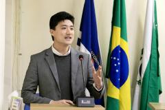 #ParaQuemNaoPodeVer foto de um homem jovem, de pele clara e cabelo preto, vestindo terno cinza, suéter escuro e camisa branca. Ele está em pé atrás de um púlpito, falando ao microfone, com expressão sorridente. Atrás dele, há bandeiras, incluindo a do Brasil e outra azul com a palavra "NITOINI" parcialmente visível. O ambiente parece ser formal, possivelmente uma apresentação ou discurso.