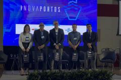 #ParaQuemNaoPodeVer Na imagem do evento, há cinco pessoas (quatro homens e uma mulher) em pé, posando para foto em um palco. Todos estão vestidos de forma formal, com crachás pendurados no pescoço. Eles seguram sacolas pretas com o logo do evento. Ao fundo, há um painel azul grande com a palavra "INOVAPORTOS" escrita na vertical. Também é possível ver algumas cadeiras vazias no palco e plantas decorativas na frente. O ambiente parece ser de uma conferência ou seminário.