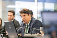 #praquemnaopodever foto mostra homem de terno azul, camisa branca e gravata verde sentado olhando para além da câmera e falando ao microfone. Ao fundo, estão desfocados os colegas que prestam atenção ao homem moreno e olhar centrado.