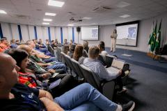 #ParaQuemNaoPodeVer Auditório com várias pessoas sentadas em cadeiras, assistindo a uma apresentação. No fundo da sala, uma mulher está em pé, falando ao microfone, com um telão atrás dela exibindo um slide. Há bandeiras do Brasil e de outros estados ou instituições ao lado do palco. As pessoas na plateia parecem estar prestando atenção, algumas com crachás e blocos de anotações. O ambiente é iluminado por luzes de teto e tem paredes brancas com detalhes em azul.