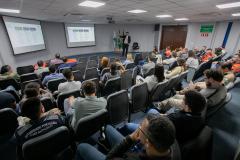 #ParaQuemNaoPodeVer Sala de aula ou auditório com várias pessoas sentadas em cadeiras, todas voltadas para a frente, assistindo a uma apresentação. Na frente da sala, há uma pessoa de pé, aparentemente palestrando, e dois telões com slides projetados na parede. No canto direito, há três bandeiras, incluindo a do Brasil. Muitas pessoas estão usando camisetas cinza com a inscrição “AUTORIDADE PORTUÁRIA PORT AUTHORITY” nas costas. O ambiente parece formal e voltado para treinamento ou reunião.
