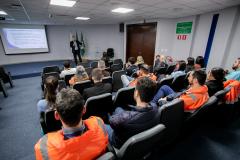 #ParaQuemNaoPodeVer Sala de reuniões vista de trás, com várias pessoas sentadas em cadeiras, assistindo a uma apresentação. Muitas dessas pessoas estão usando uniformes laranja com faixas refletivas, típicos de trabalhadores de áreas industriais ou de segurança. Na frente da sala, há uma pessoa em pé, aparentemente apresentando um slide projetado na parede. Ao lado do apresentador, estão duas bandeiras (provavelmente do Brasil e do estado). A sala tem piso azul, paredes claras e teto com iluminação embutida