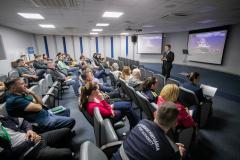 #ParaQuemNaoPodeVer Auditório com várias pessoas sentadas em cadeiras, todas de cabeça baixa. Na frente, um homem de terno está em pé, também de cabeça baixa, próximo a um púlpito transparente. Ao fundo, há dois telões projetando a mesma imagem, com fundo roxo e texto branco, mas o texto não está totalmente legível na foto. O ambiente é fechado, com iluminação de teto e paredes claras.