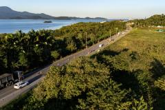 #ParaQuemNaoPodeVer Foto aérea de uma estrada rodeada por vegetação densa e árvores. À esquerda da imagem, há um caminhão e um carro branco na estrada. À direita, é possível ver um corpo d’água azul (provavelmente um rio ou mar) e montanhas ao fundo. A estrada corta a paisagem diagonalmente, e há postes de energia acompanhando o trajeto. 