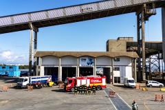 #ParaQuemNaoPodeVer  foto de um pátio industrial ou de uma garagem de veículos grandes. No centro da imagem, há dois caminhões: um caminhão de bombeiros vermelho com a inscrição "BOMBEIROS" e outro caminhão branco com detalhes azuis, que parece ser de resgate ou suporte. Ambos estão estacionados próximos a um prédio de estrutura robusta, com cobertura alta e colunas de concreto. Há cones e barreiras laranja ao redor dos veículos, indicando uma área isolada. Algumas pessoas com coletes refletores estão próxi