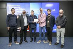 #ParaQuemNaoPodeVer Foto de seis homens posando juntos em frente a um painel azul. Eles estão em pé, lado a lado, sorrindo para a câmera. Um dos homens, no centro, segura um objeto retangular, que parece ser um prêmio ou uma placa. O painel atrás deles tem textos em português, incluindo frases como “PARTICIPE DO EVENTO”, “Segurança e Responsabilidade no Armazenagem”, e “Sistema Profissional”. Todos estão vestidos de maneira casual ou social, com camisas, blazers e calças.