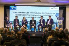 #ParaQuemNaoPodeVer Foto de um painel em um evento corporativo, com cinco pessoas (quatro homens e uma mulher) sentadas no palco, participando de um debate. No fundo, há um grande telão com o texto: "Logística no Brasil ONDE ESTAMOS HOJE E COMO ALCANÇAR OS INVESTIMENTOS NECESSÁRIOS PARA O DESENVOLVIMENTO ECONÔMICO SUSTENTÁVEL DA REGIÃO SUL". O público está sentado de frente para o palco, acompanhando a discussão. No canto inferior direito da tela, há um púlpito transparente com a mesma identidade visual do 