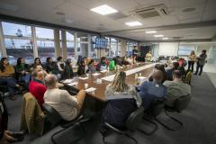 #ParaQuemNaoPodeVer Foto mostra uma reunião em uma sala de conferências. Ao redor de uma mesa grande e retangular, há muitas pessoas sentadas, a maioria prestando atenção em alguém que está falando ou apresentando (próximo a um quadro de projeção na ponta da mesa). A sala tem janelas grandes ao fundo, por onde é possível ver navios. As pessoas na reunião são de perfis variados e algumas estão com laptops ou papéis na mesa. A imagem sugere um ambiente corporativo ou de trabalho colaborativo. 
