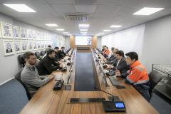 #ParaQuemNaoPodeVer Sala de reuniões com uma mesa longa de madeira, onde diversas pessoas estão sentadas dos dois lados, aparentemente participando de uma reunião. Algumas pessoas usam uniformes laranja e cinza, que parecem ser de alguma empresa ou serviço. Há microfones e pequenos dispositivos em frente a cada pessoa na mesa, sugerindo que é uma reunião formal ou corporativa. Na parede à direita estão pendurados vários quadros com fotos de pessoas, provavelmente fotos de ex-diretores, presidentes ou homena