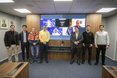 #ParaQuemNaoPodeVer Foto de sete homens em uma sala de reunião corporativa. Eles estão todos em pé, lado a lado, olhando para a câmera. Quatro deles estão vestidos de forma casual e três usam roupas mais formais (terno ou camisa social). Dois dos homens ao centro estão utilizando uniformes: um de jaqueta laranja e outro de jaqueta amarela, que parecem ser de alguma empresa ou serviço. Atrás deles, há uma televisão grande na parede exibindo uma imagem colorida com pessoas em uma chamada de vídeo, sugerindo q