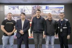 #ParaQuemNaoPode Ver Imagem mostra cinco homens posando para uma foto em grupo. Eles estão em pé, lado a lado, com um fundo que parece ser uma imagem de uma área industrial ou de construção, com estruturas metálicas e guindastes. Um dos homens, na parte superior esquerda, veste uma camiseta preta com a palavra "Mormaii" escrita. Outro homem, ao lado dele, está de terno azul escuro. Os demais usam roupas casuais ou jaquetas, e um deles, na parte inferior, usa um crachá e uniforme preto com detalhes 